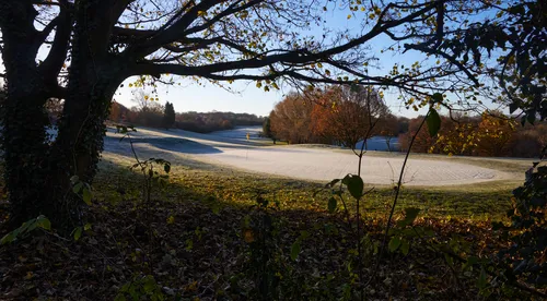 This landscape photograph shows a frozen golf course at Tapton Park in Chesterfield, United Kingdom, viewed from underneath trees on the edge of the park. The scene captures a frosty winter morning, with the sun low in the sky casting long shadows and illuminating the park’s grass with a layer of frost. The trees frame the golf course, many of which have lost their leaves, underlining the winter season. The area is part of Tapton Park, known for its natural surroundings and recreational spaces, and the photograph highlights the golf course as the main subject, surrounded by nature and mature trees. The setting gives a clear indication of the winter climate in this part of the United Kingdom.